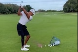 A golfer stands on a driving range with golf balls at his feet, taking a swing with a wedge.