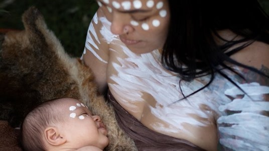 Aboriginal woman covered in white body paint holds and looks down at a child.