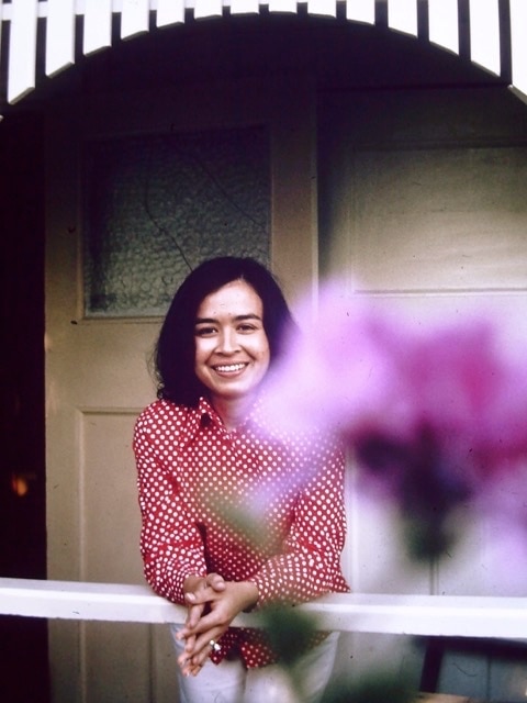 A Malaysian woman wearing a red shirt poses for a photo on a verandah, in front of a pink flower