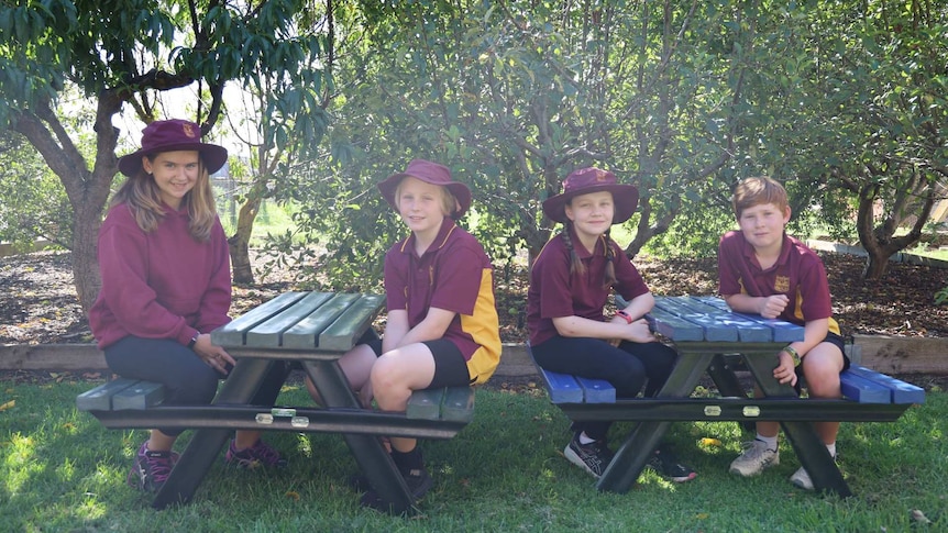 Four kids in maroon and yellow uniforms sit smiling at two green and blue picnic tables