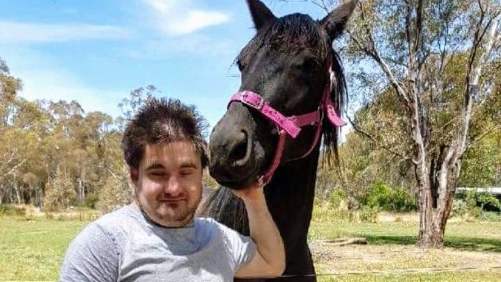 A young man stands next to a black horse with a pink bridle.