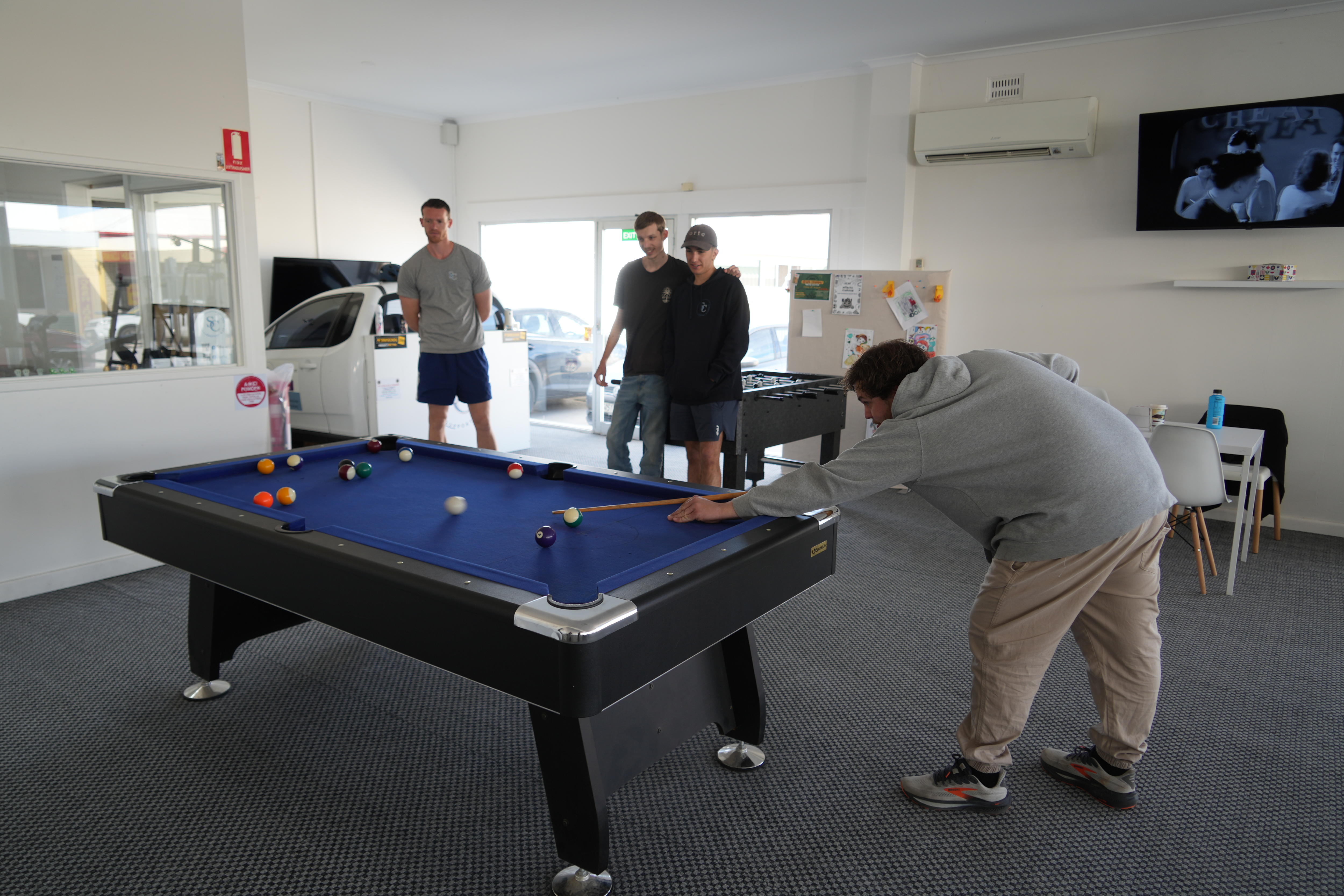 A group of men gathered around a pool table in a building, one in the process of hitting a ball