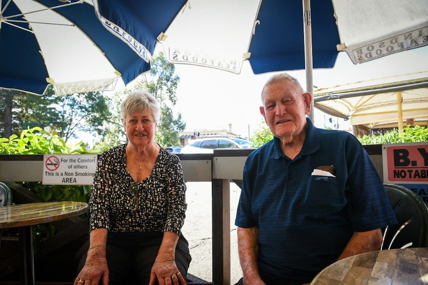 An elderly man and woman seated at a metal table smile at the camera. 