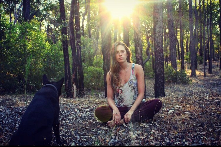 A young woman sits crossed legged in the bush with a black dog looking at her.