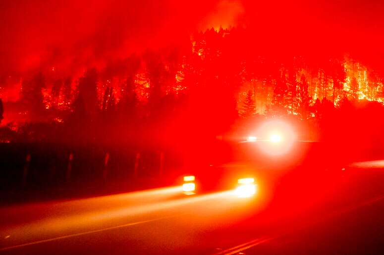 You view a smoke-covered, blood-red sky at night with emergency vehicles racing past the camera with their headlights on.