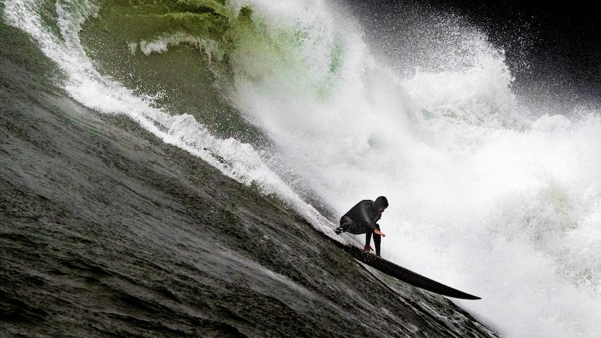 Huge 3 story wave dark green water surfer is half way down the massive face wearing full wetsuit boots and hood in cold water