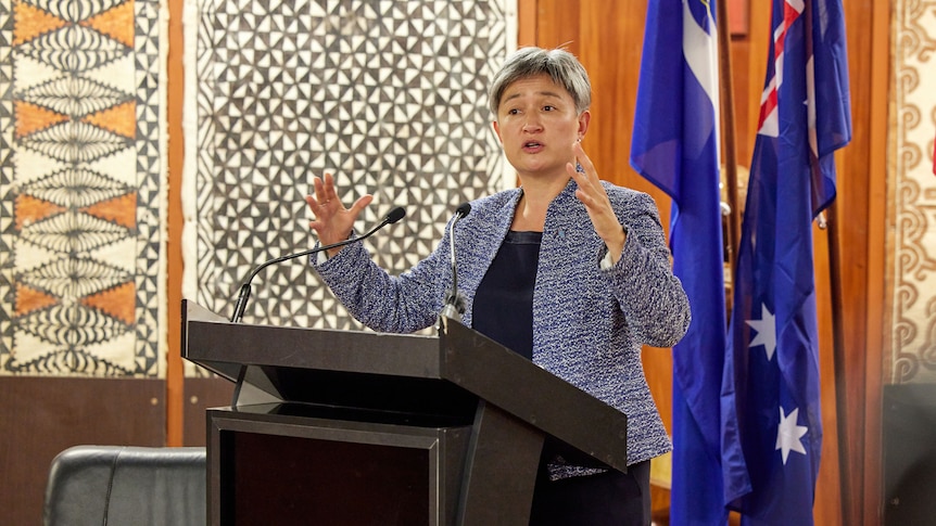 Penny Wong speaking behind a lectern in front of the Australian flag. 