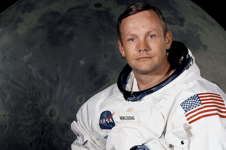 Neil Armstrong in his space suit. Behind him is a large photograph of the lunar surface.