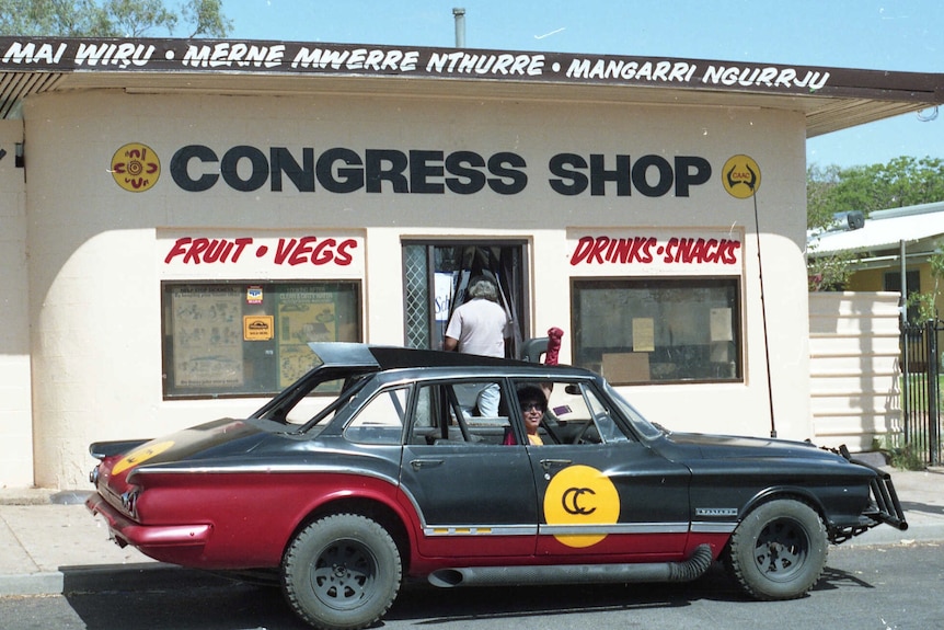 A man sits inside an old Valiant car painted in black, red and yellow.
