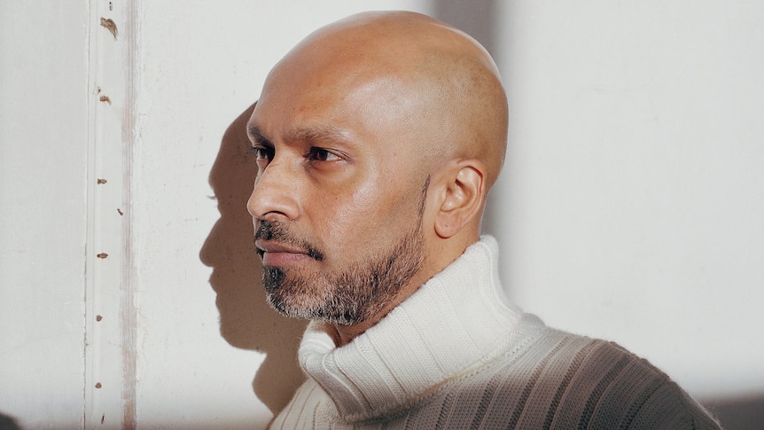 Akram Khan stands expressionless in a white room. His head is in a patch of bright light, his body is in shadow.