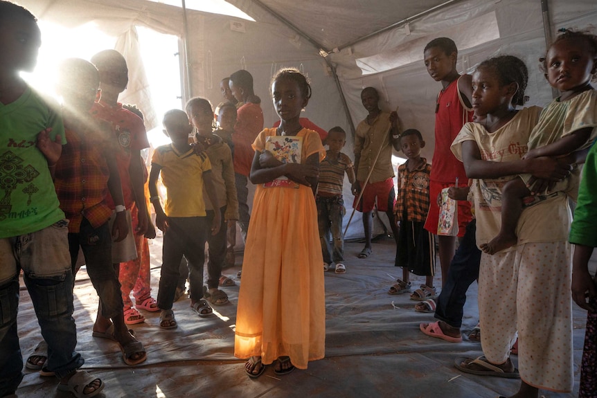 A girl in an orange dress clutching a notebook to her chest, surrounded by other children in a white tent.