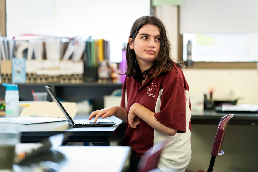 A wide shot of a teenage girl in a classroom wearing a school uniform at her computer. Her left hand is smaller than her right.