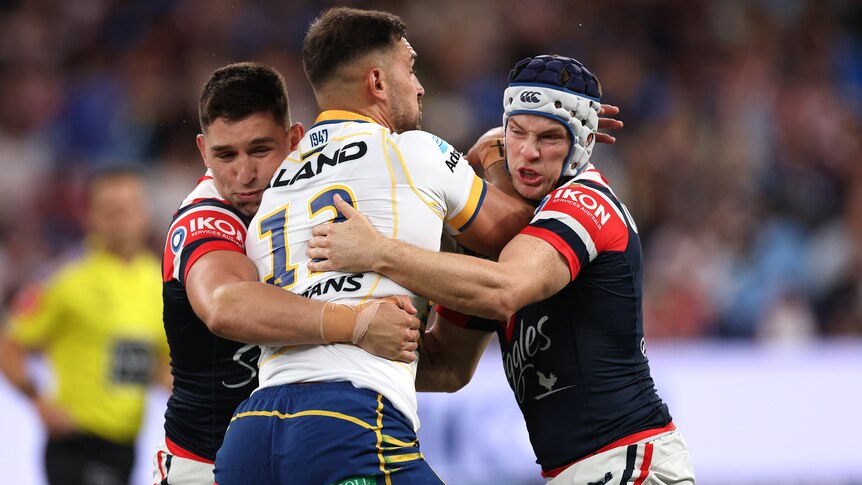 A Parramatta NRL player holds the ball as he is tackled by two Sydney Roosters opponent.
