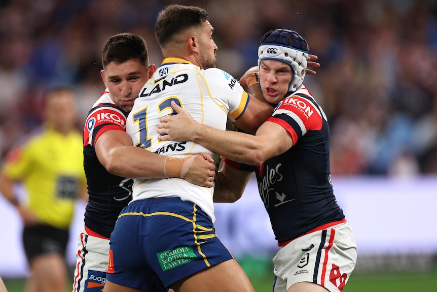 A Parramatta NRL player holds the ball as he is tackled by two Sydney Roosters opponent.