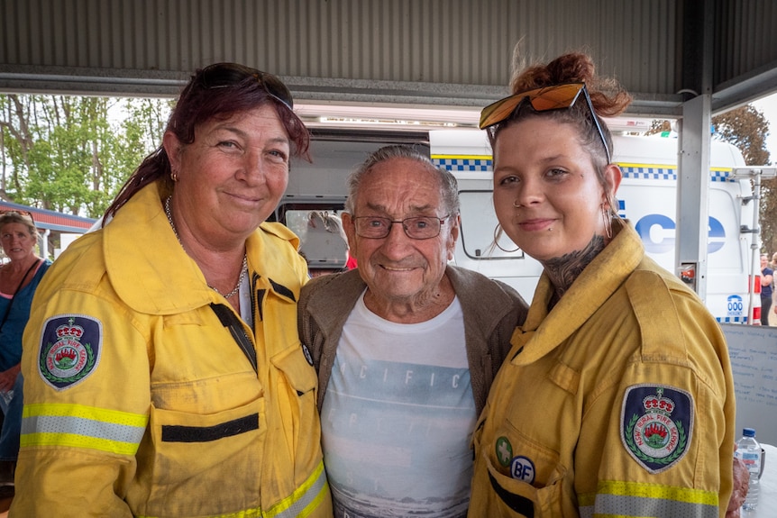 Two women stand either side of an elderly man. The woman on right is in her early 20s and has neck tattoos.