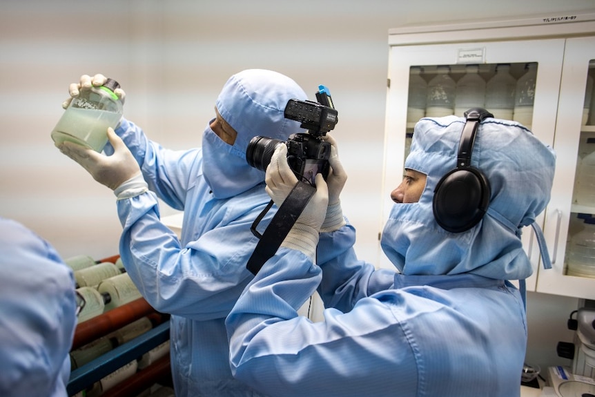 Edgerton in surgical scrubs holding jar with white cloudy liquid up to camera being held by woman in scrubs.