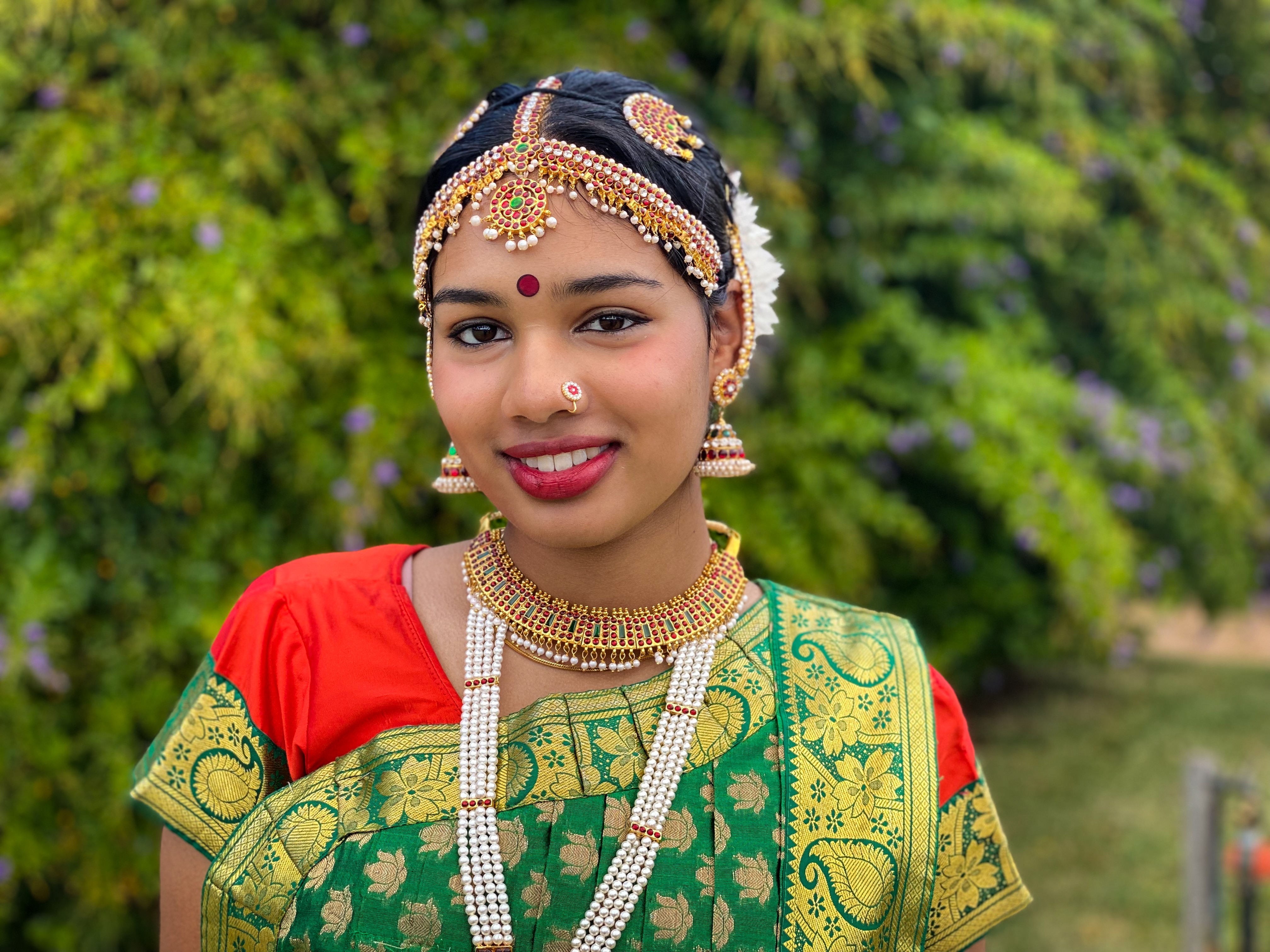 A young brown girl is adorned with gold, beaded jewellery and colourful sari. She is smiling confidently.
