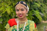 A young brown girl is adorned with gold, beaded jewellery and colourful sari. She is smiling confidently.