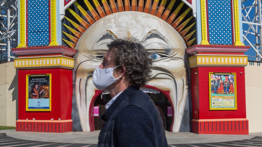 A person wearing a cloth face mask walks past Melbourne's Luna Park on a sunny day.