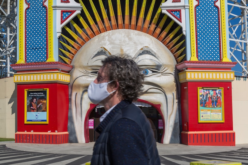 A person wearing a cloth face mask walks past Melbourne's Luna Park on a sunny day.