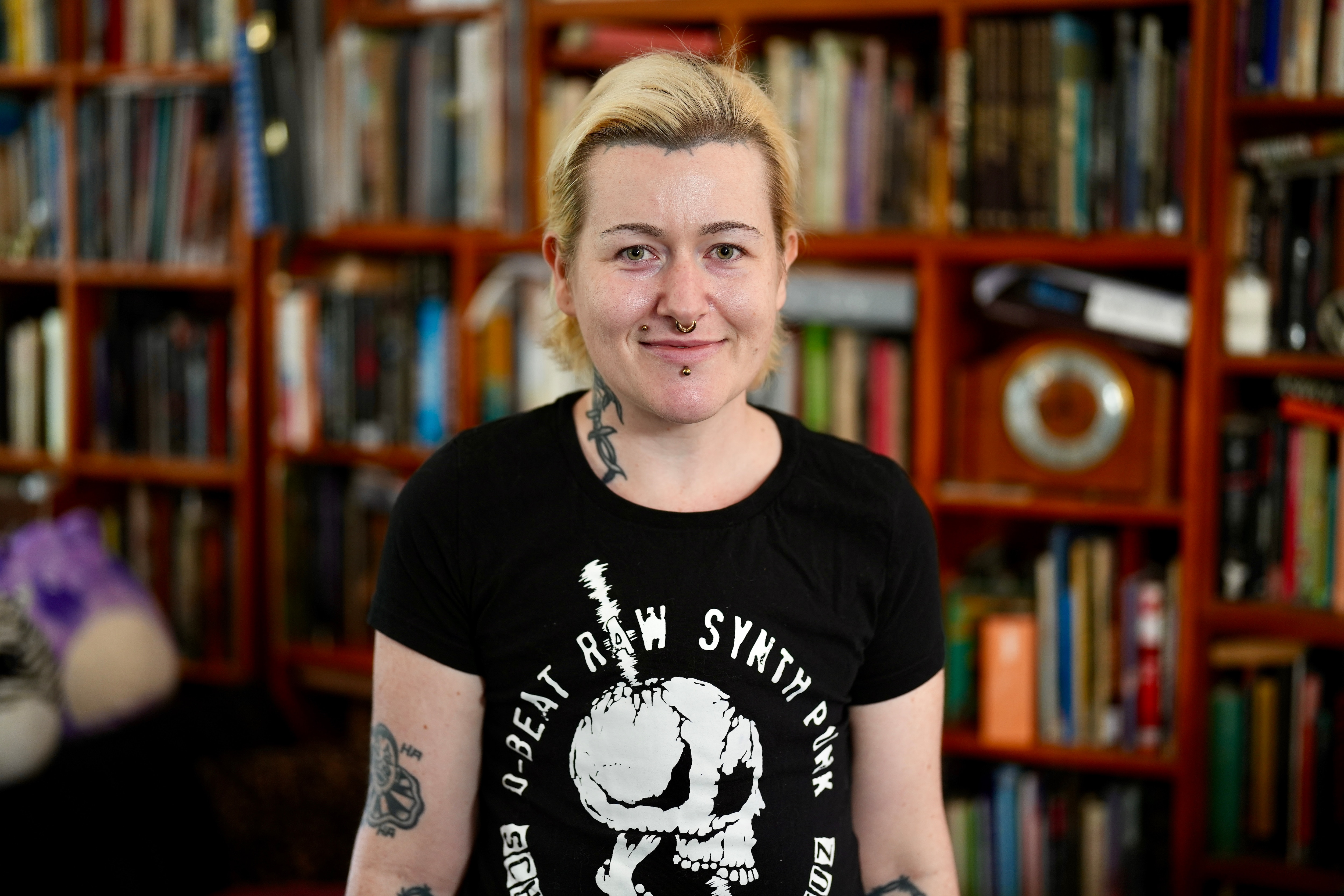 DJ Lana Zilla smiling in a black and white t-shirt, with tattoos on their neck and arms and blonde dyed hair.