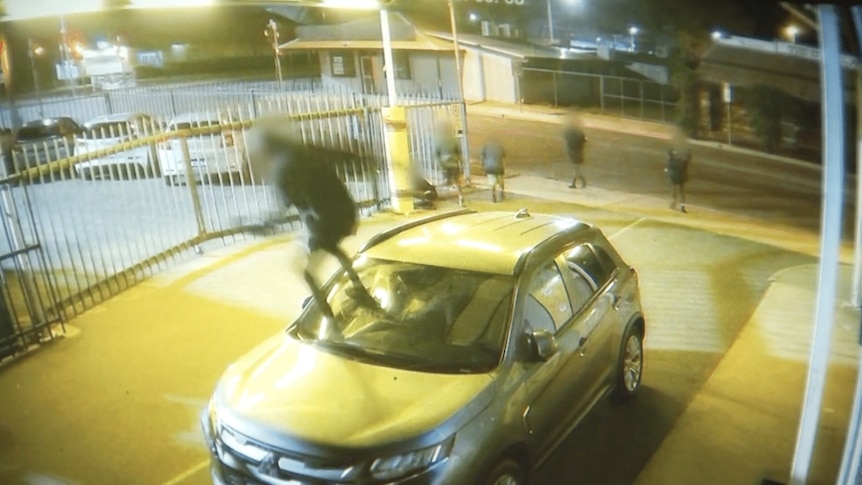A screenshot of CCTV of someone smashing a car windscreen with their foot.