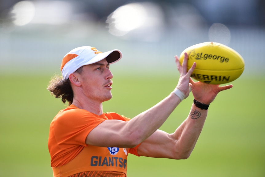 A man in an ornage shirt with a cap on catches an AFL ball.