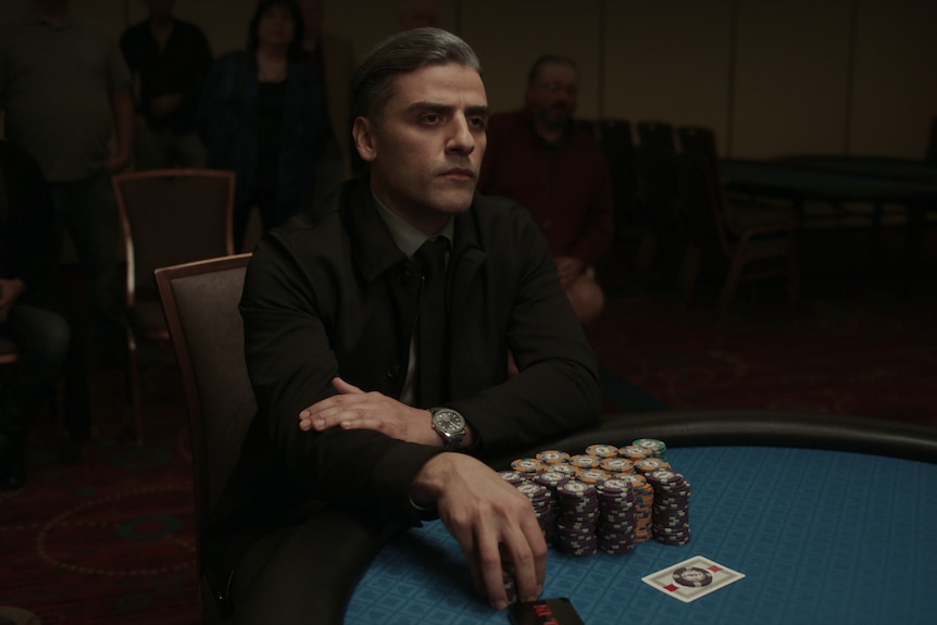A man in his early 40s with greying hair slicked back sits a gaming table, a stack of chips in front of him