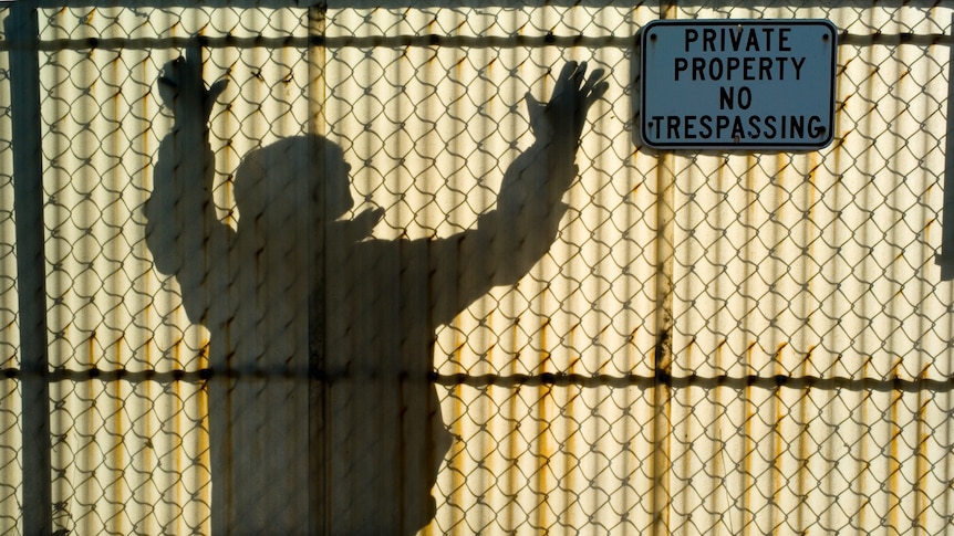 Man reaches for the top of a translucent fiberglass  fence while a sign on the other side warns of not trespassing.