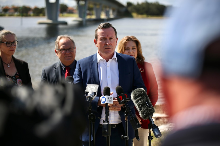 A man in a white shirt and blue jacket speaking at a press conference in front of a body of water.