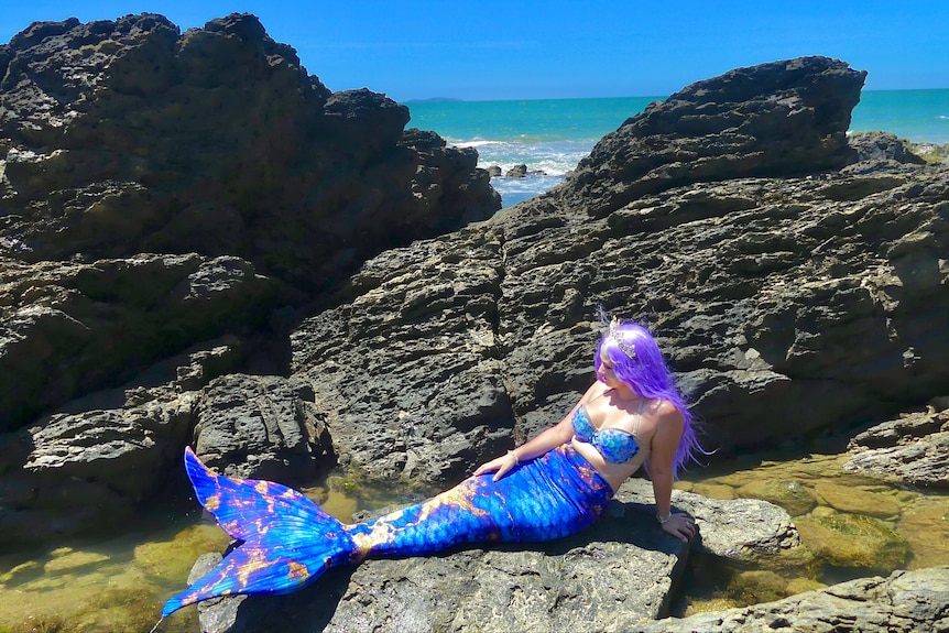 A woman wearing a purple wig, bra and a mermaid tail on some coastal rocks at a beach