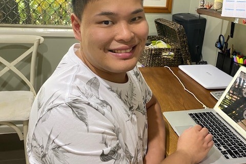 A young Asian man sits at a computer at a desk and looks to the camera, smiling. He wears a white and grey shirt.