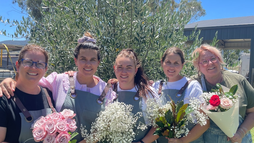 Four women holding various flowers in front of an olive tree smiling. 