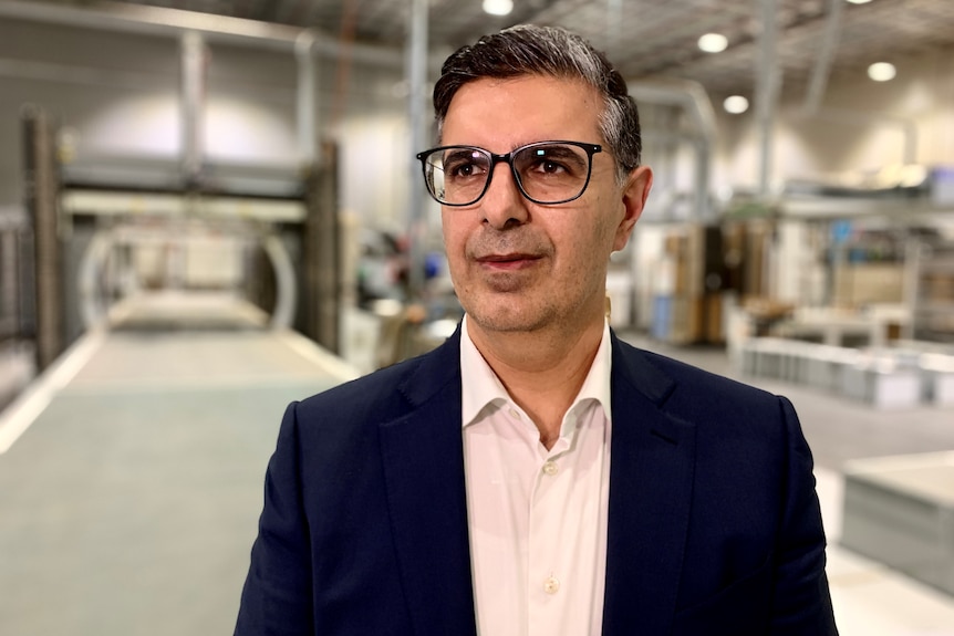 A middle aged man wearing glasses and a dark suit jacket over a light shirt stands in a kitchen manufacturing factory.