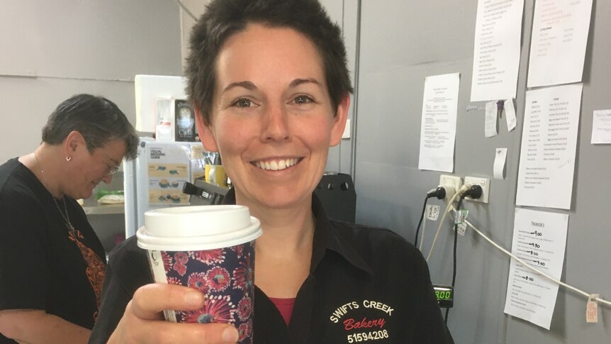 A smiling woman with short, dark hair holds up a coffee.