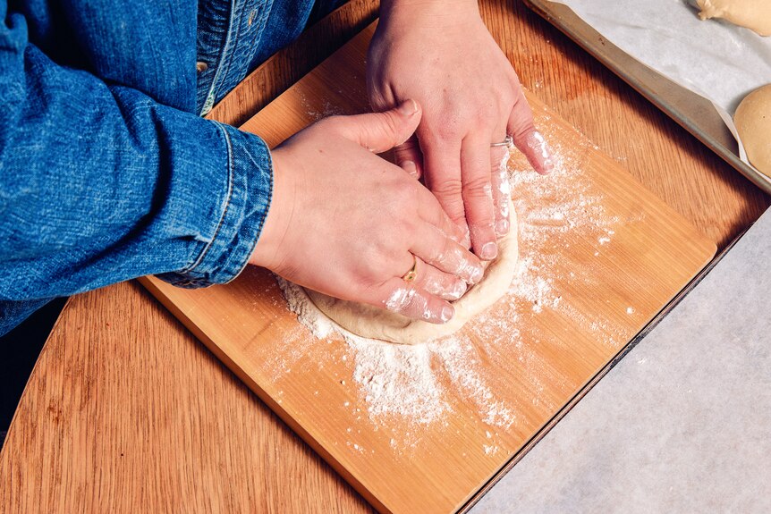 Two hands knead a ball of dough into a flat pizzette shape on a wooden board covered in flour.