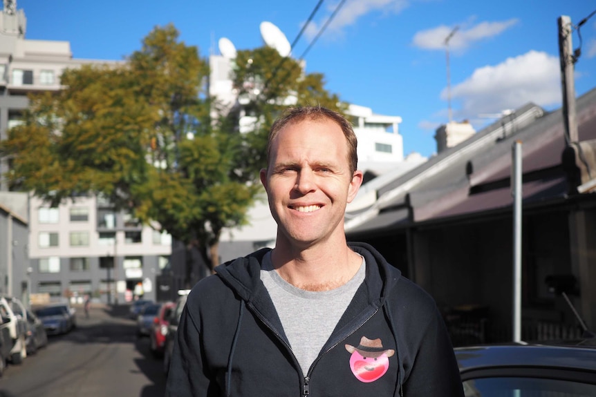 A man wearing a black hoodie smiles while squinting into the sun, standing on a quiet car-lined street.