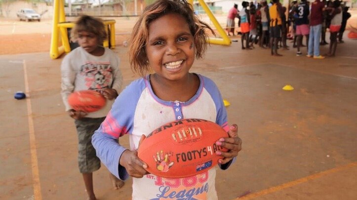 A smiling Aboriginal girl holds an AFL football