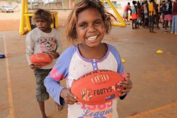 A smiling Aboriginal girl holds an AFL football