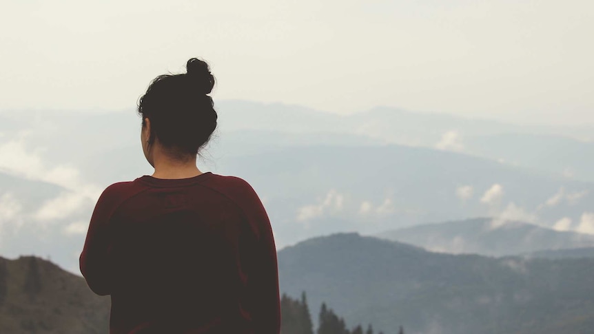 A woman stands alone in contemplation, looking out at snow capped mountains.