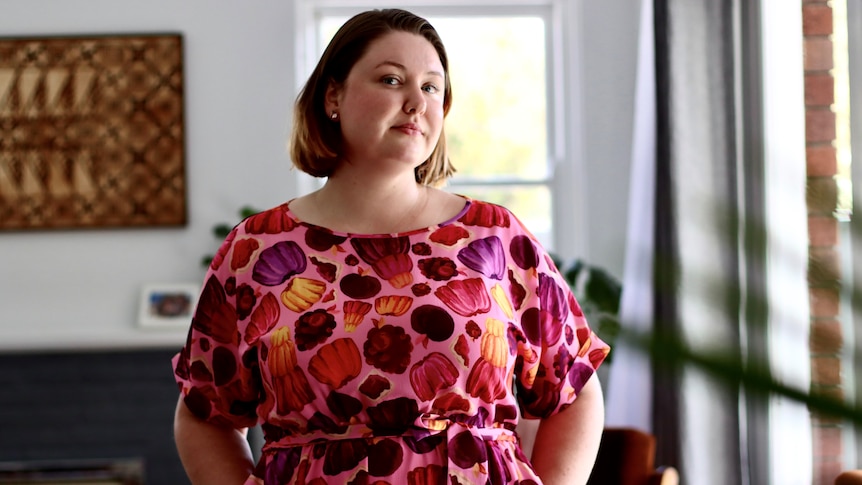 A woman with short brown hair wears a brightly coloured dress in her living room. She's staring straight at the camera.