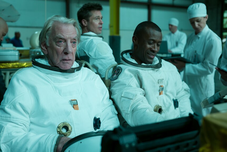 Interior shot of three men in space suits, no helmets, seated looking ahead.