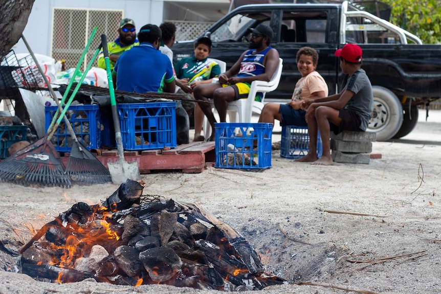 Men and young children sitting around a fire outside in front of a ute.