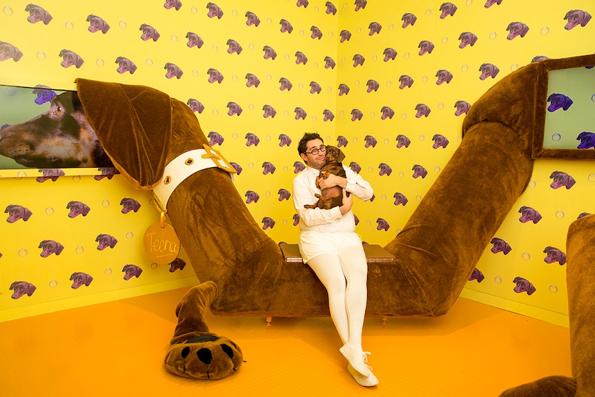 Colour photo of artist David Capra posing with his pet Daschund Teena, in front of a large scale version of Teena and wallpaper.