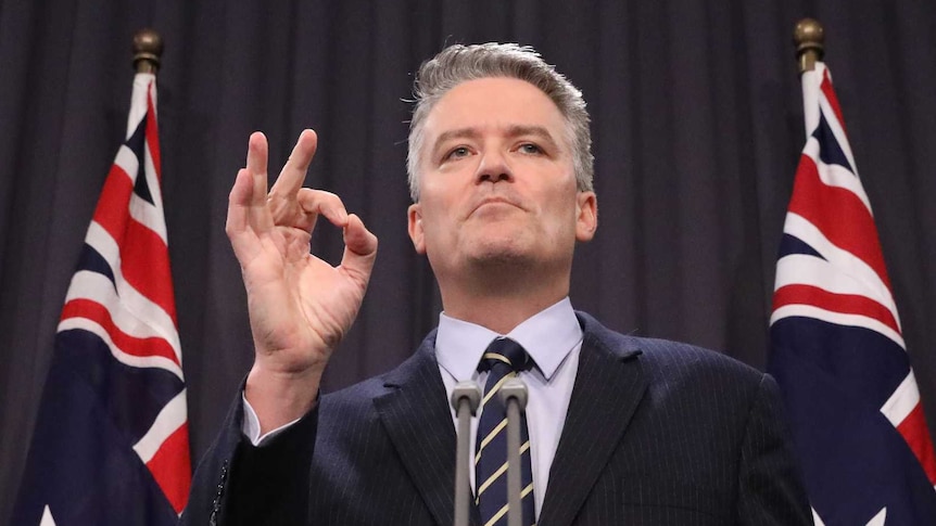 Mathias Cormann holds up his right hand in an 'OK' motion, while standing at a lectern in front of two Australian flags.