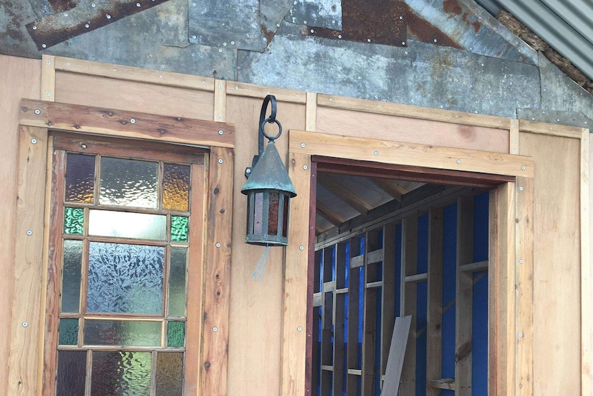 A small, bare wooden unfinished structure, with corrugated iron roofing, rustic lamp and small stained-glass window.