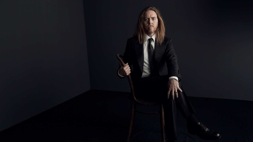 Tim Minchin sitting on a chair in front of a grey backdrop.