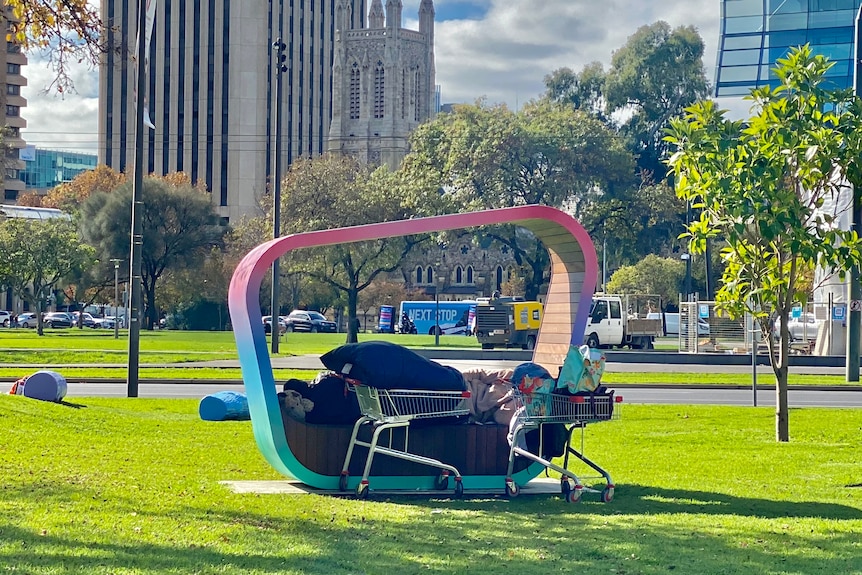 A person sleeps under blankets on a colourful art installation in a green square with a church in the background