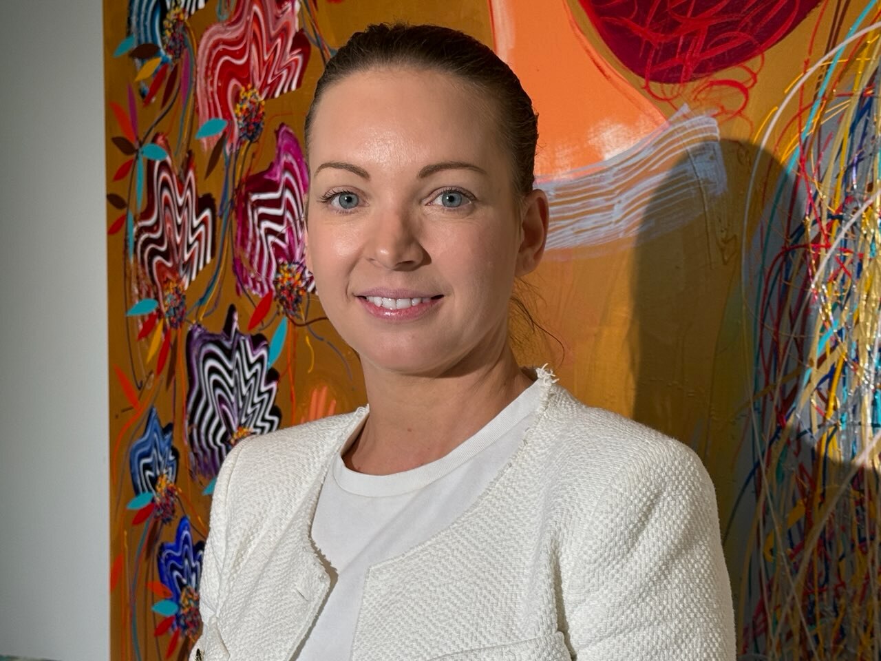 A white woman brown hair in a bun, wearning a white blazer standing in front of an orange painting.