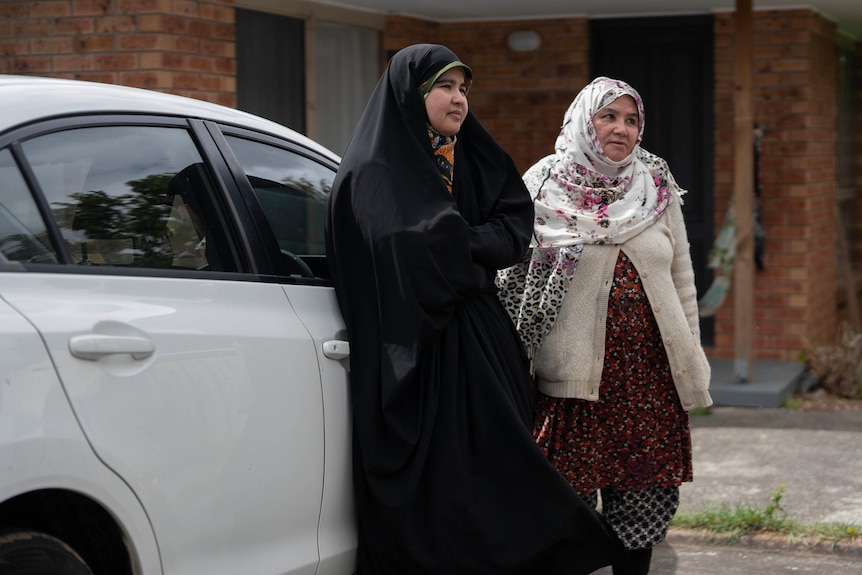 Two Hazara women in traditional clothing stand next to a parked car.